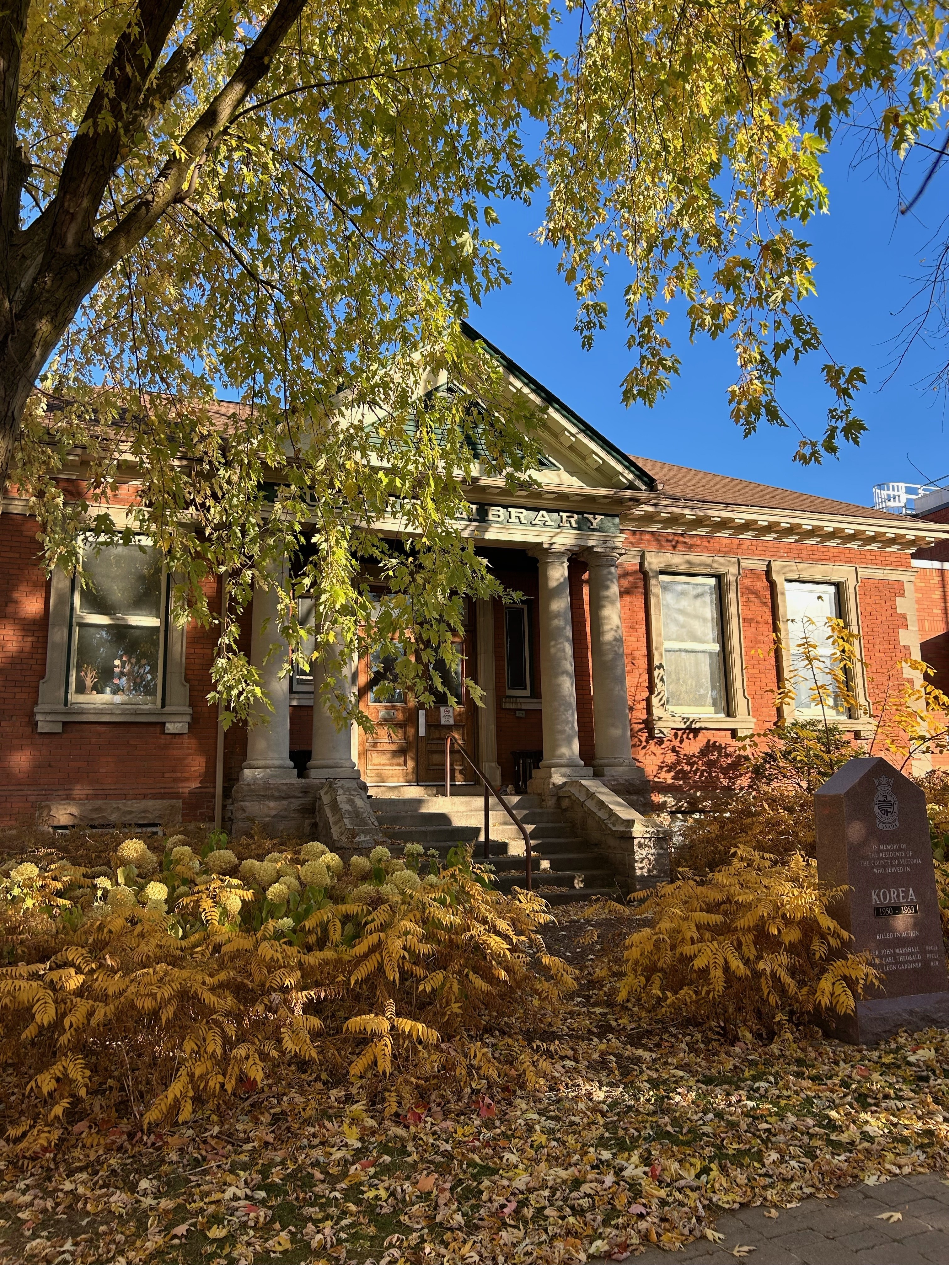 Historic brick library with white columns, surrounded by autumn foliage. Sunlight filters through trees, casting a warm, inviting glow.